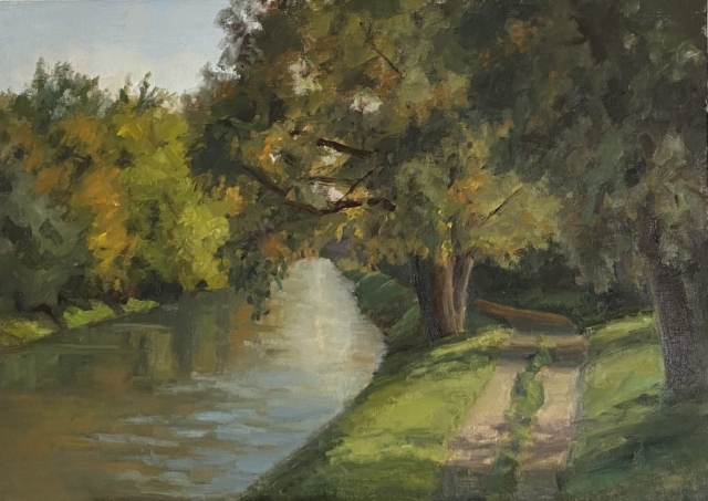Path leads along the river. Willow trees grow along and cast shadows on the path.